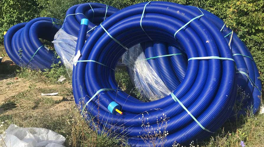 Zwijane rury preizolowane WATTS Microflex z karbowaną niebieską osłoną zewnętrzną, zabezpieczone taśmami i przechowywane na zewnątrz przed instalacją.