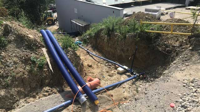 Instalacja preizolowanych rur WATTS Microflex w wykopie na placu budowy, z odsłoniętymi połączeniami rurociągu i pobliskim budynkiem przemysłowym.