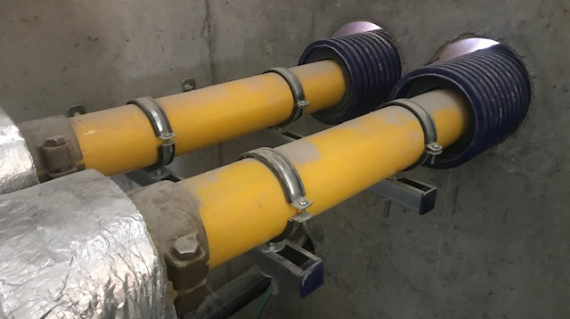 Preizolowane rury WATTS Microflex z żółtymi rurami wewnętrznymi i niebieską karbowaną osłoną zewnętrzną, bezpiecznie zamontowane na wspornikach ściennych i przechodzące przez betonową ścianę.