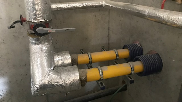 Preizolowane rury WATTS Microflex z żółtymi rurami wewnętrznymi i niebieską karbowaną osłoną zewnętrzną, bezpiecznie zamontowane na wspornikach ściennych i podłączone do izolowanego systemu zaworów w pomieszczeniu gospodarczym o betonowych ścianach.