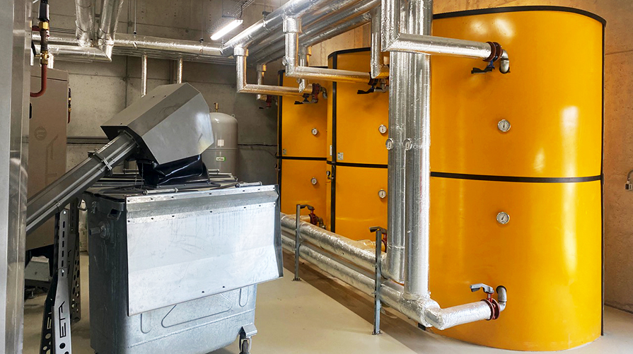 Kotłownia z dużymi żółtymi zbiornikami termicznymi połączonymi z izolowanym systemem rur i kotłem na biomasę ze zautomatyzowanym systemem podawania paliwa, zintegrowanym z preizolowanymi rurami WATTS Microflex dla wydajnej dystrybucji ciepła.