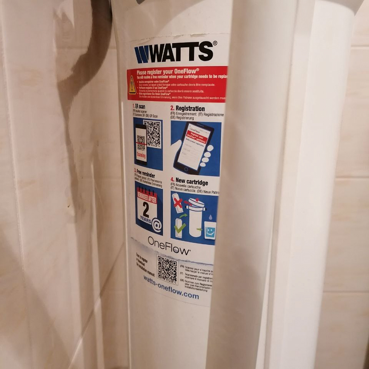 Zbliżenie systemu filtracji wody WATTS OneFlow ze szczegółową etykietą przedstawiającą instrukcje instalacji, rejestrację kodu QR i wytyczne dotyczące wymiany wkładu.