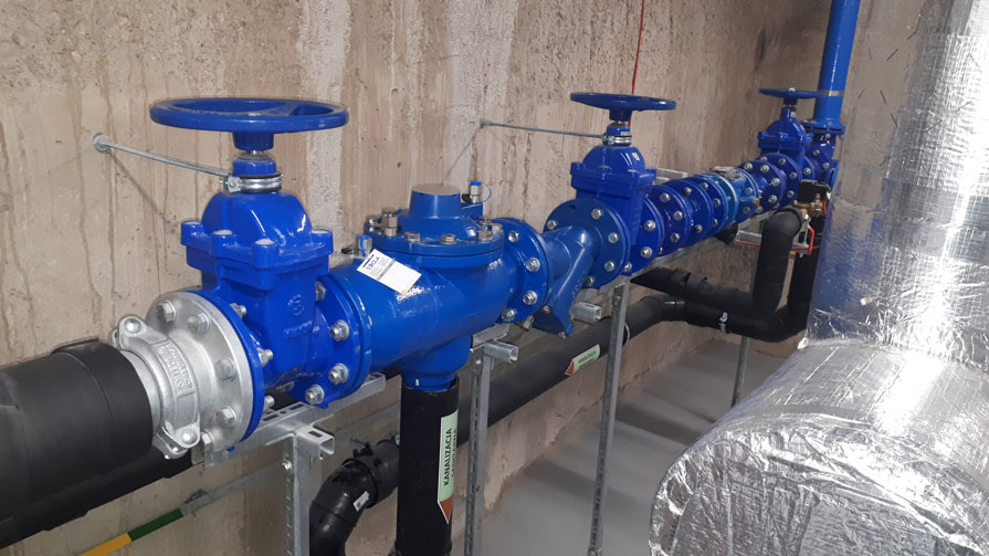 System zapobiegający przepływowi zwrotnemu WATTS BA4760 DN100 z niebieskimi zasuwami i połączeniami kołnierzowymi, zainstalowany na metalowej konstrukcji wsporczej w pomieszczeniu mechanicznym.