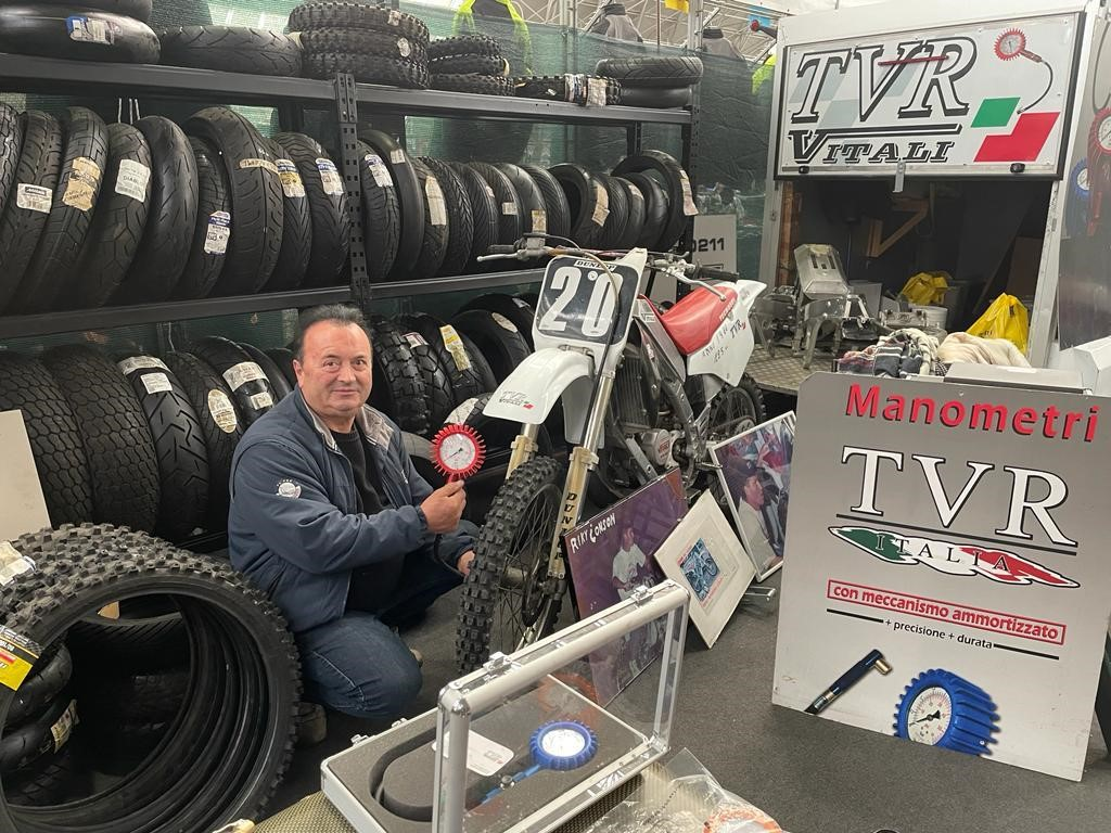 Hombre presentando un manómetro WATTS junto a una moto de motocross de época, con estanterías de neumáticos de moto al fondo.
