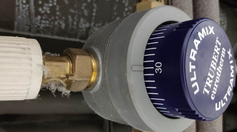 Термостатичний змішувальний клапан ULTRAMIX від TRUBERT Eurotherm, встановлений у фітнес-клубі Calypso для підтримки безпечної температури гарячої води. Великий план регулятора з налаштуванням на 30°C і латунним з'єднанням із трубопроводом.