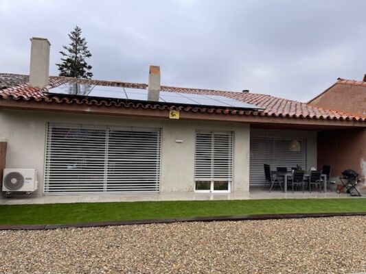 Casa a un piano a Barcellona con tetto in tegole di terracotta, pannelli solari installati, grandi porte scorrevoli in vetro con persiane, una zona pranzo all'aperto e un cortile in ghiaia con erba sintetica.