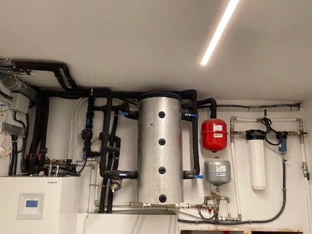 Boiler room in a house in Barcelona, featuring a central cylindrical insulated water tank, black insulated pipes, a red expansion vessel, a white filtration unit, and various plumbing and heating system components mounted on a white wall